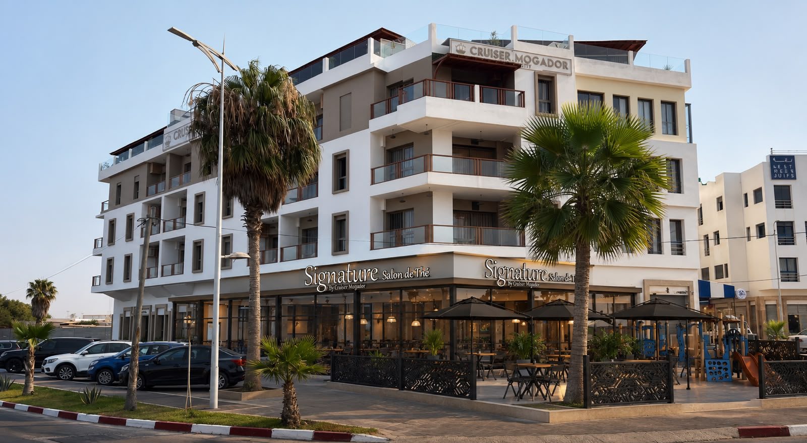 Le bâtiment vu depuis la rue : immeuble blanc de l'hôtel Cruiser Mogador, enseignes Signature sur le socle commercial, palmiers et terrasse — vue d'architecte